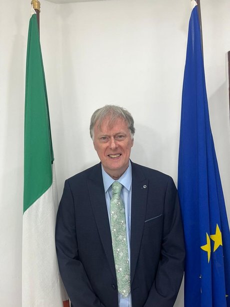 Ambassador Patrick McManus standing in front of the Irish and EU flags.